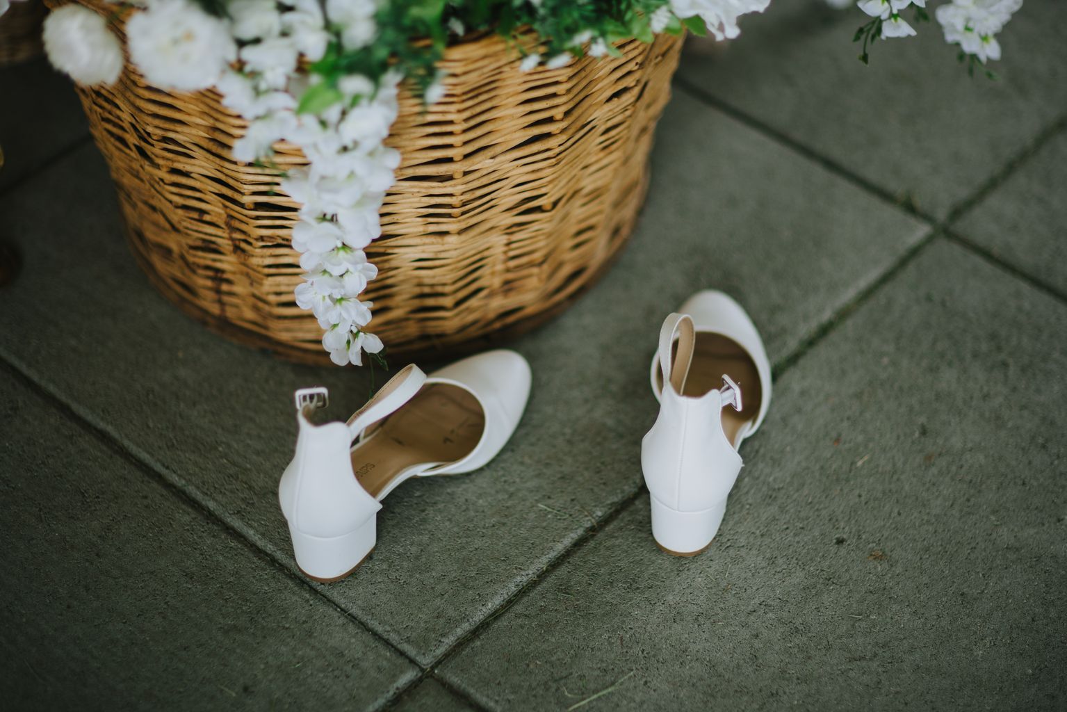 White bridal shoes and woven basket with greenery on gray stone pavement