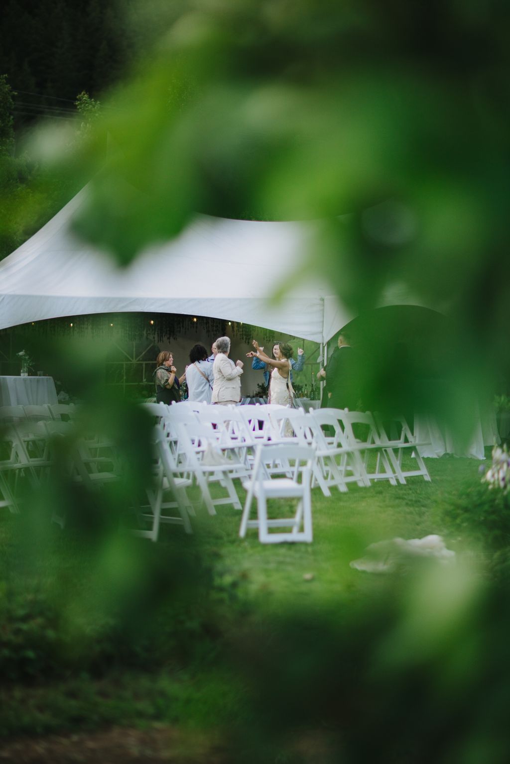 Outdoor wedding ceremony under white tent with guests seated in white chairs, viewed through soft green foliage in foreground