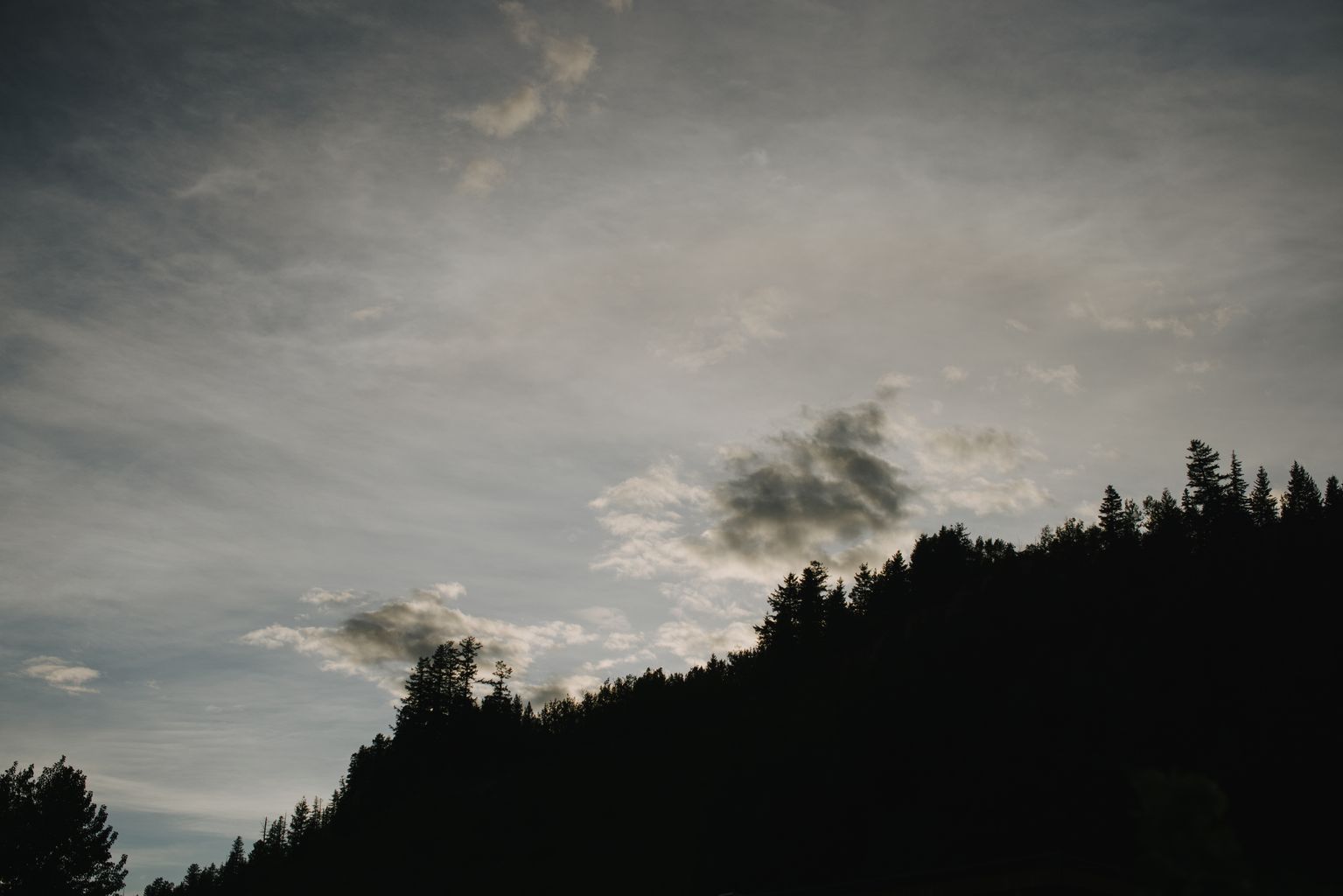 Dramatic storm clouds gather above a dark forested treeline under moody gray sky