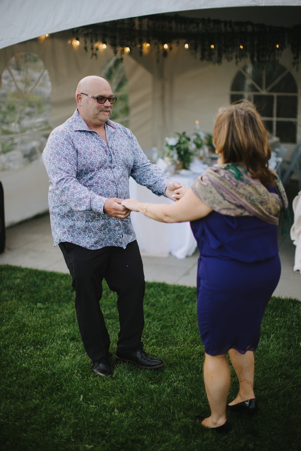 Man in patterned shirt and glasses holds hands with woman during evening outdoor reception under draped canopy