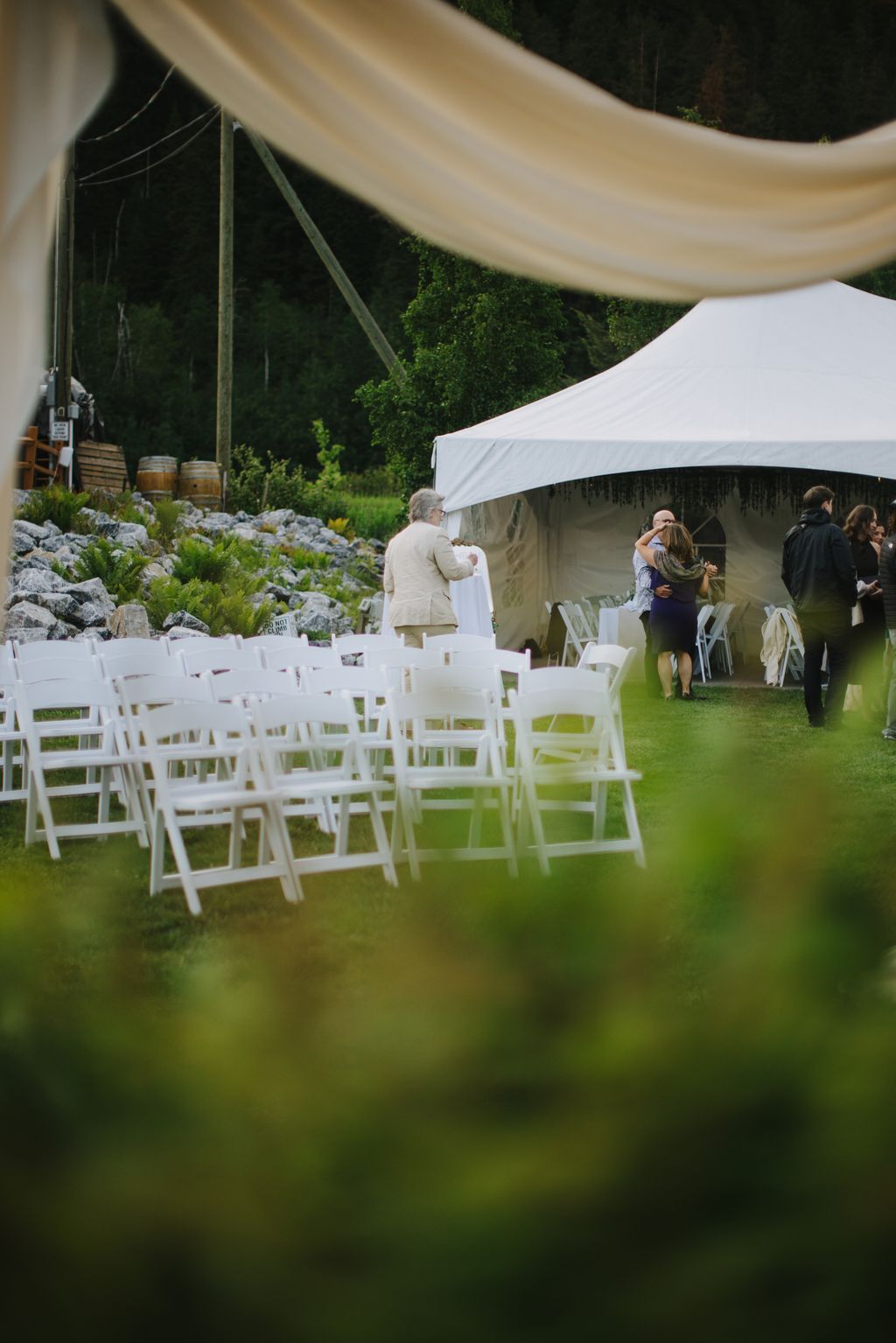 Elegant outdoor wedding ceremony with white chairs arranged on manicured lawn, beige fabric draping overhead, lush greenery backdrop, and guests in formal at…