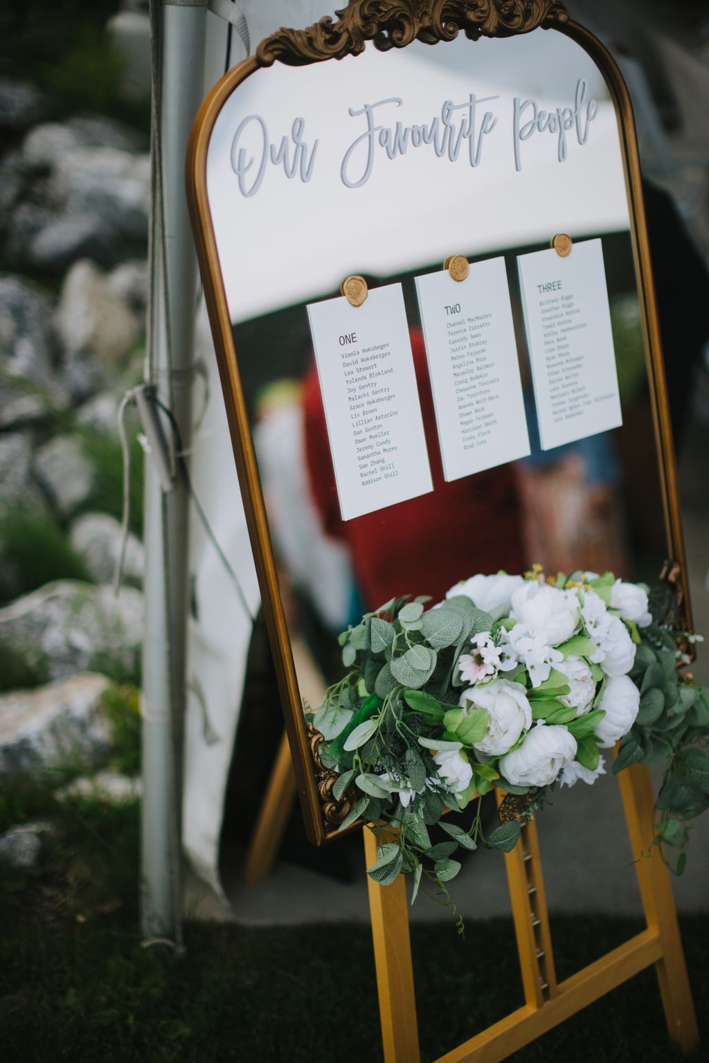Ornate gold-framed seating chart with handwritten calligraphy header, two numbered table cards, and white flower arrangement with eucalyptus greenery