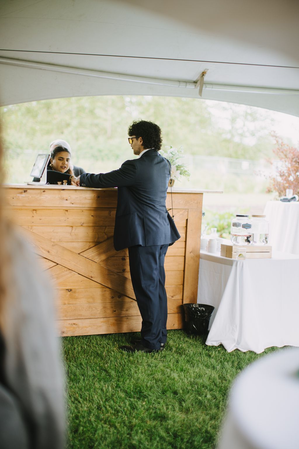 Groom in navy suit gazes out window of wooden barn venue, holding white flower, soft natural light