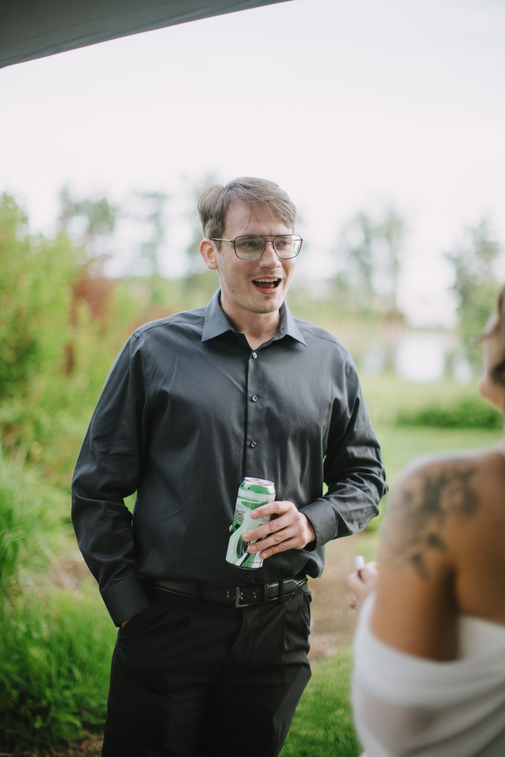 Groom in dark shirt and glasses smiles while holding a beverage outdoors with blurred green landscape behind him