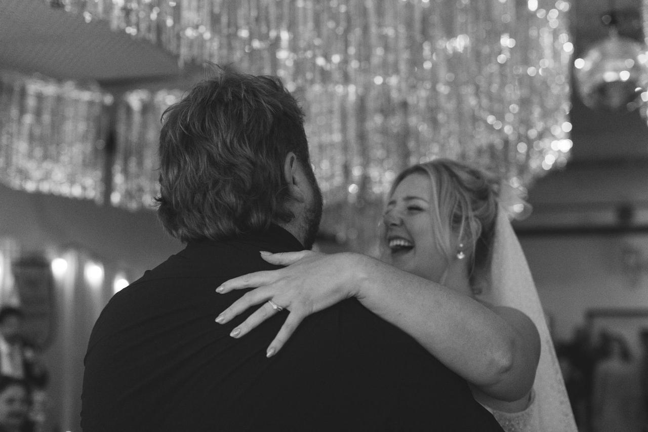 Bride and groom embrace against glowing city lights, her hand showing wedding ring, both laughing