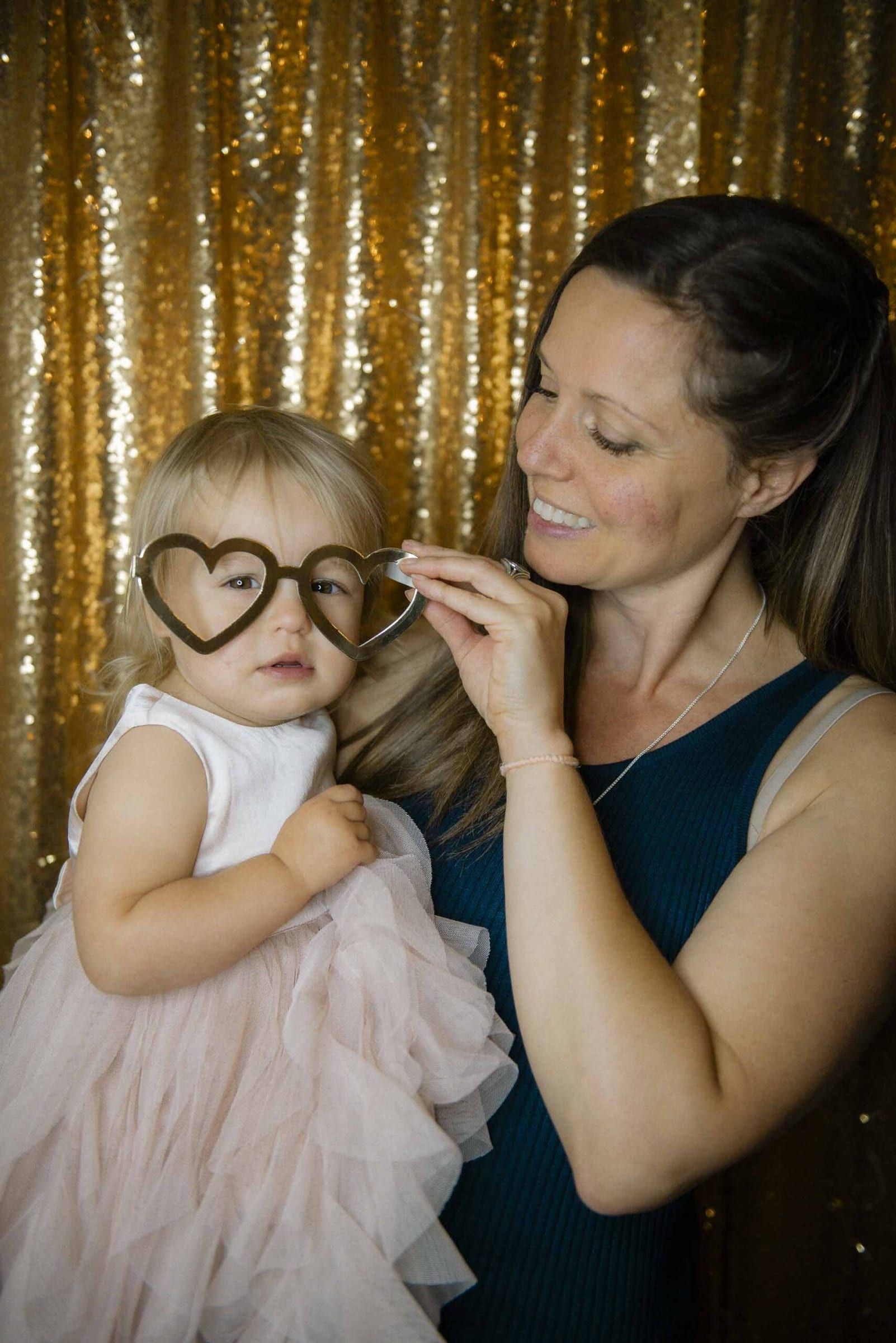 Woman and child hold a heart-shaped prop against a sequined backdrop, smiling in profile