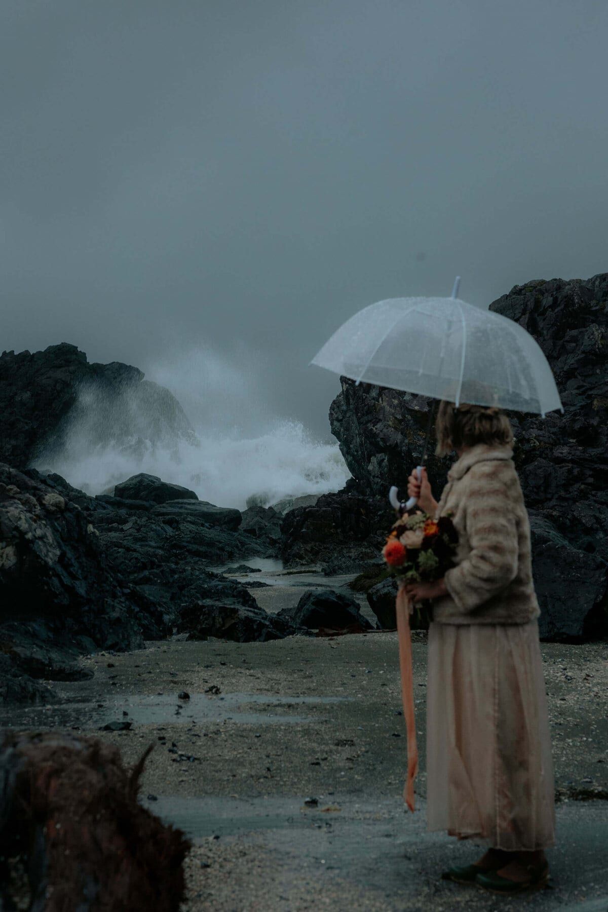 Bride under a clear umbrella watching storm waves pound the rocks