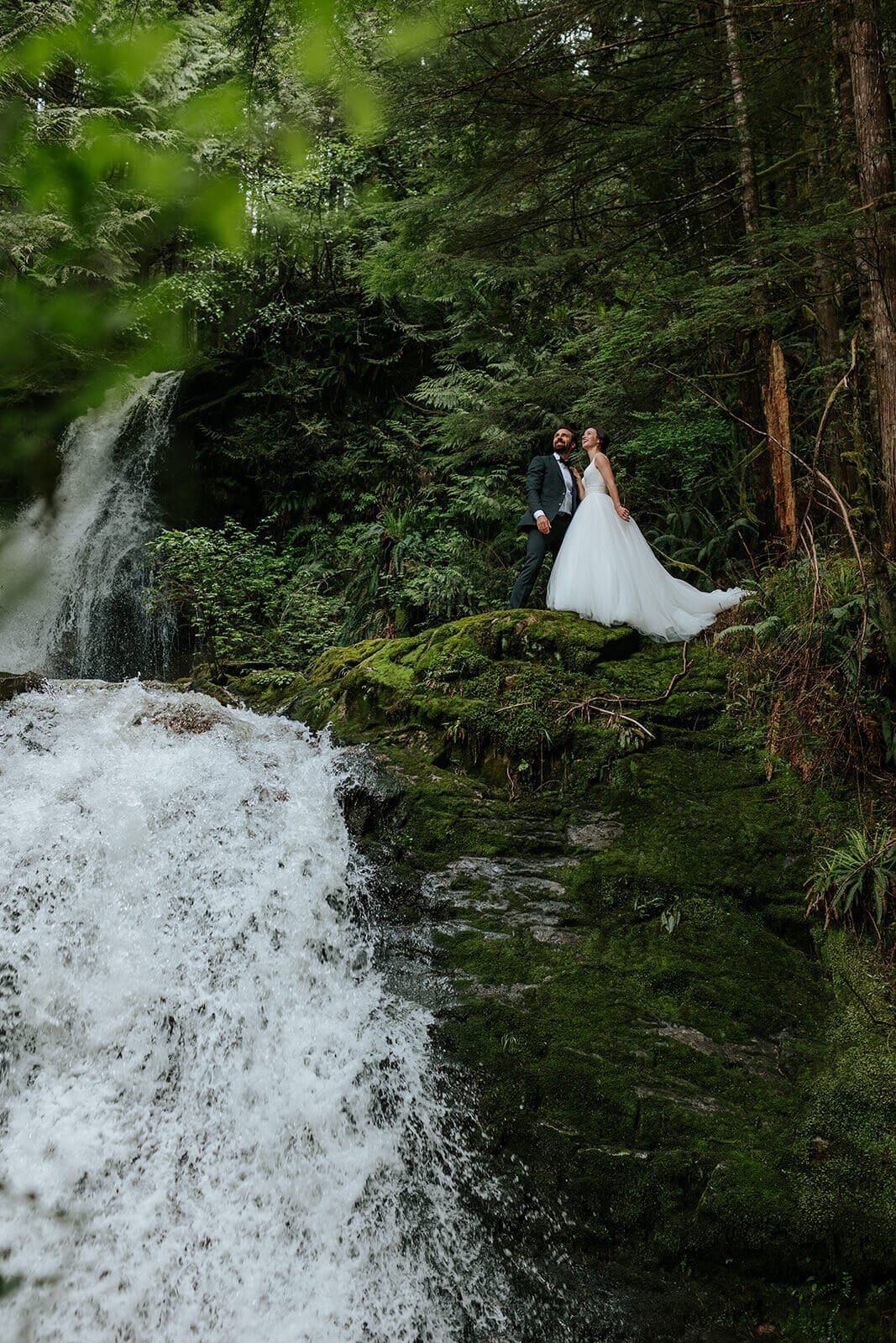 Newlyweds on a mossy ledge beside a rushing rainforest waterfall
