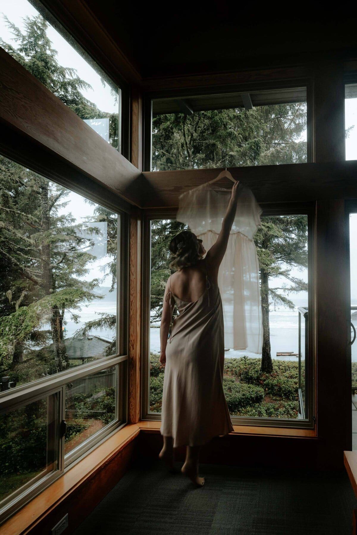 Bride reaching for her hanging dress by an ocean-view window