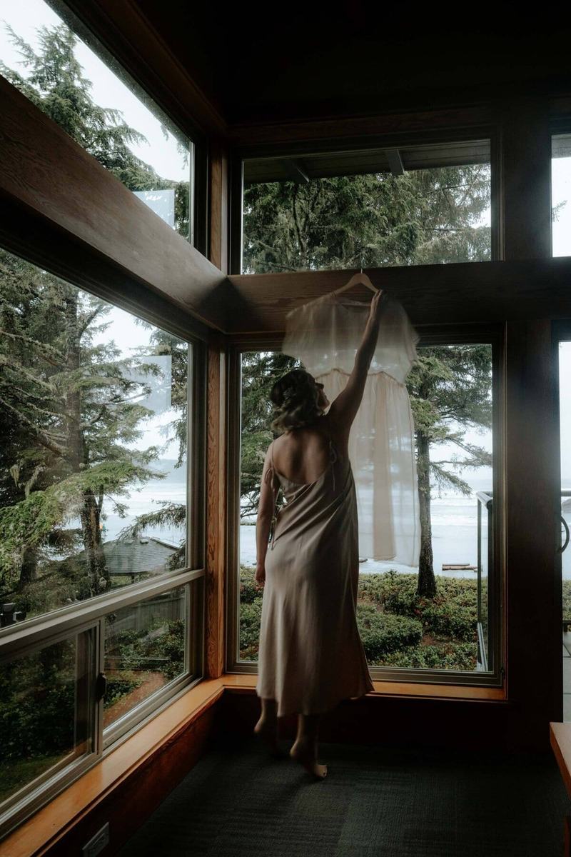Bride reaching for her hanging dress by an ocean-view window