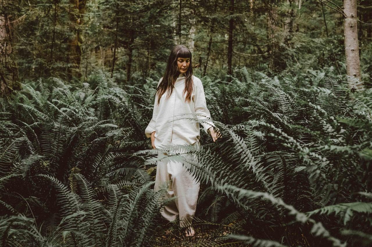 Woman in cream linen shirt and pants touches fern fronds in a mossy forest, looking down thoughtfully among green trees.