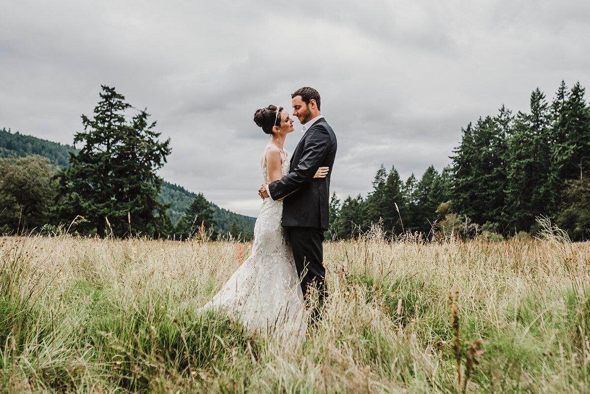 Bride and groom embracing in a tall-grass meadow beneath forested hills