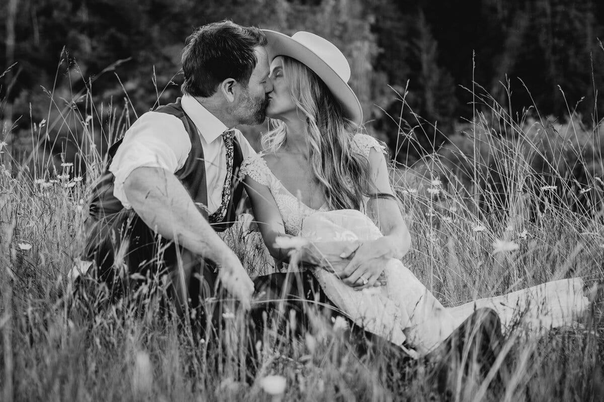 Bride in a wide-brimmed hat kissing her groom in a wildflower meadow