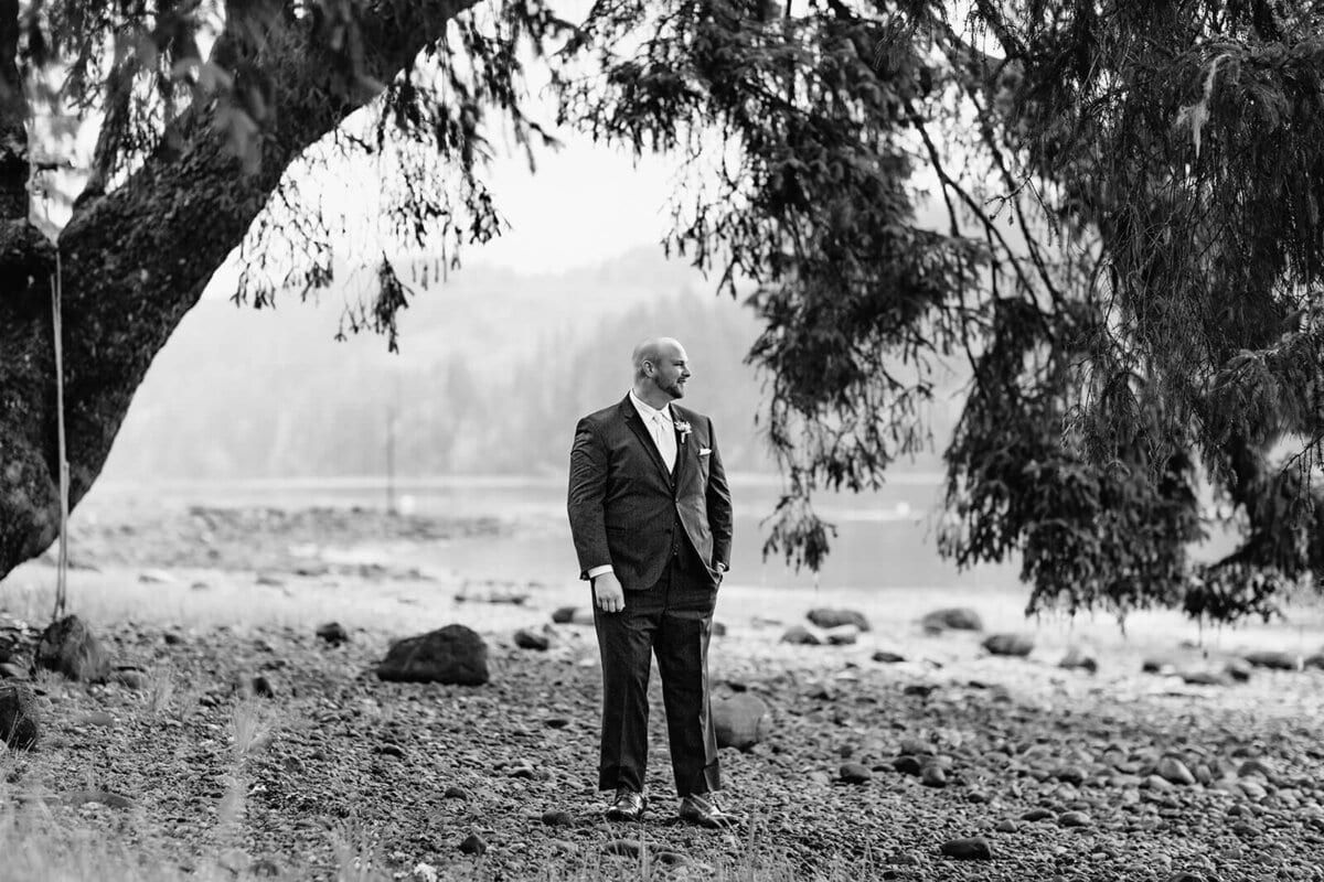 Black-and-white groom waiting beneath a sweeping cedar bough by the water