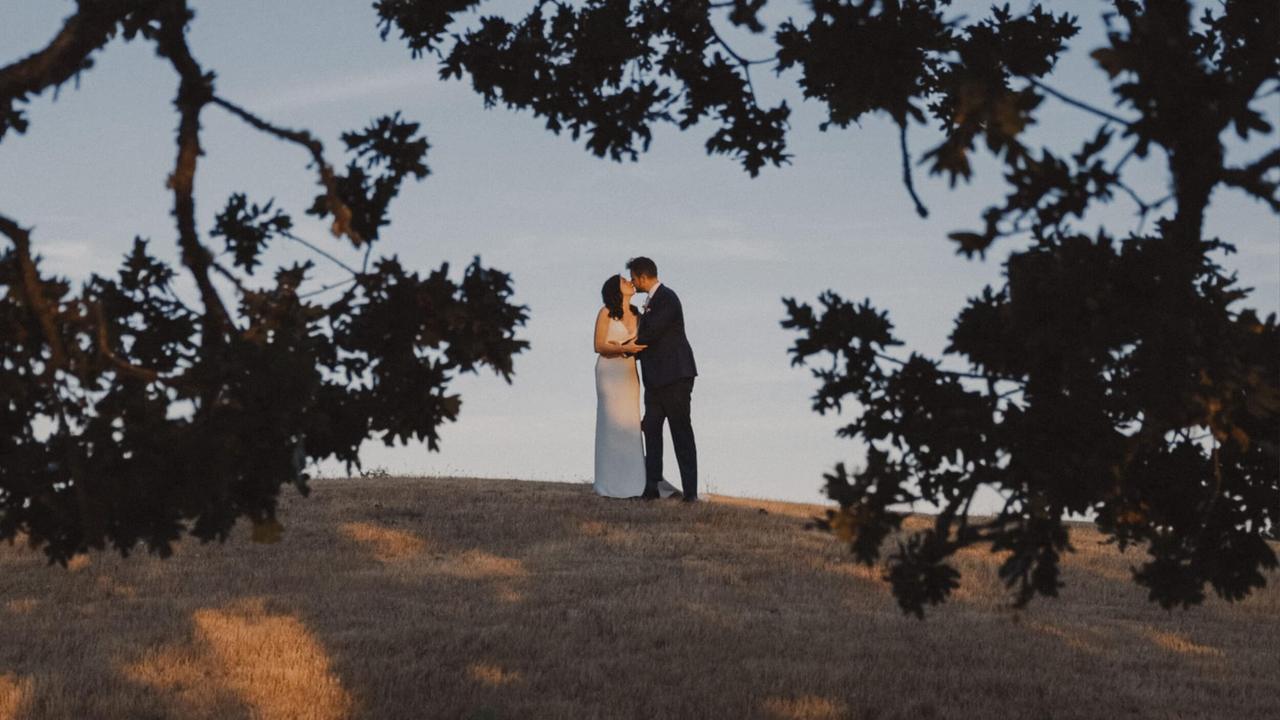 Bride and groom kiss on a grassy slope at Stonebridge Farm at dusk, framed by tree branches overhead and an overcast sky