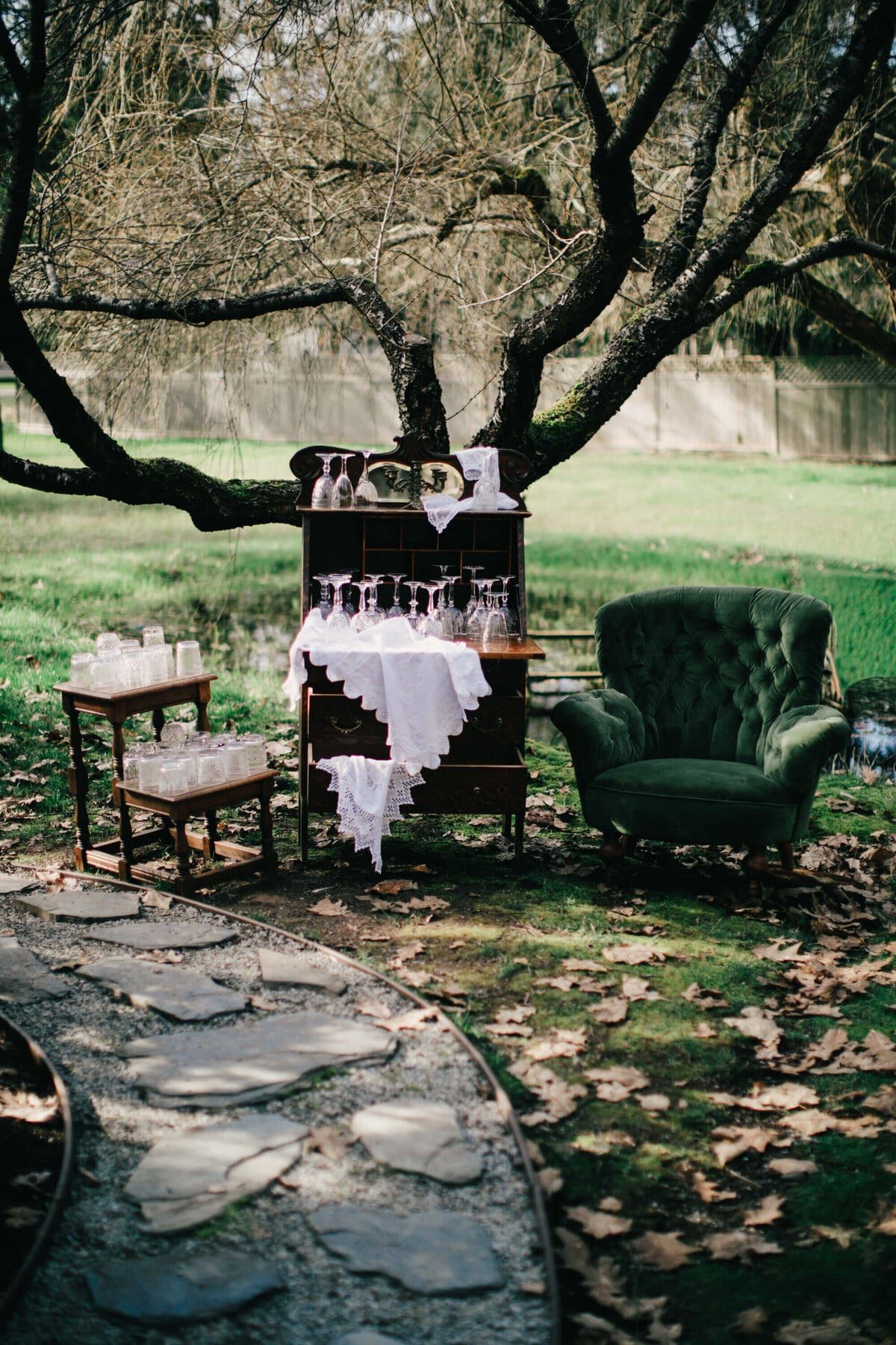 Green velvet armchair and antique glassware bar beneath a gnarled tree