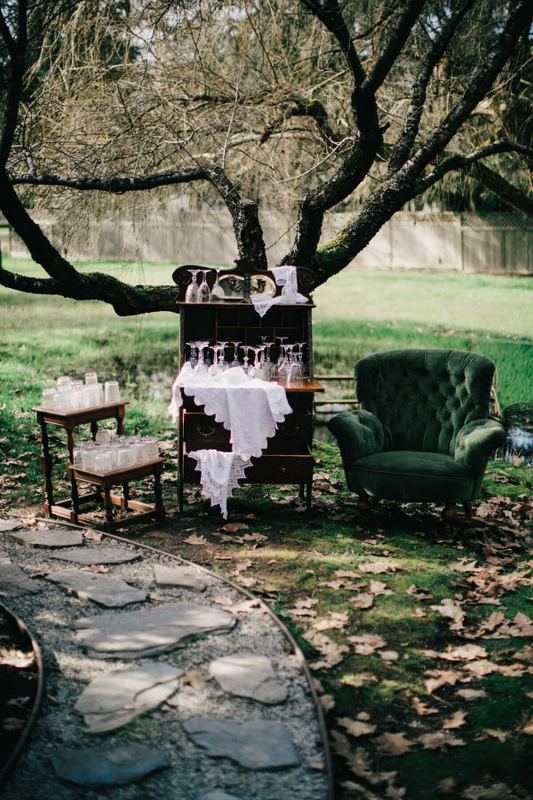 Green velvet armchair and antique glassware bar beneath a gnarled tree