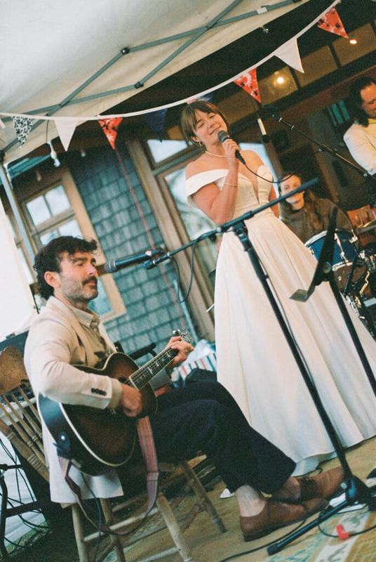 Bride singing at the microphone while a guitarist plays under the tent
