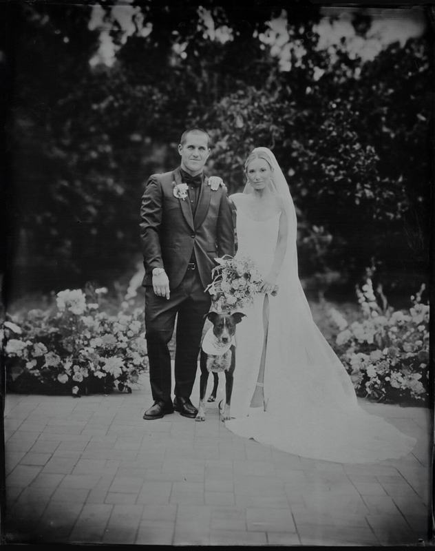 Newlyweds with their dog on a flower-lined brick path