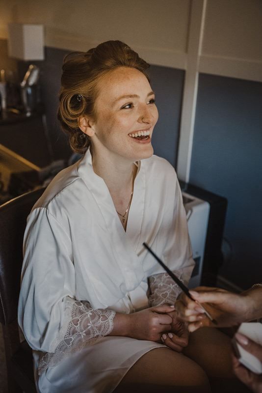 Bride in a white robe laughing while getting her makeup done
