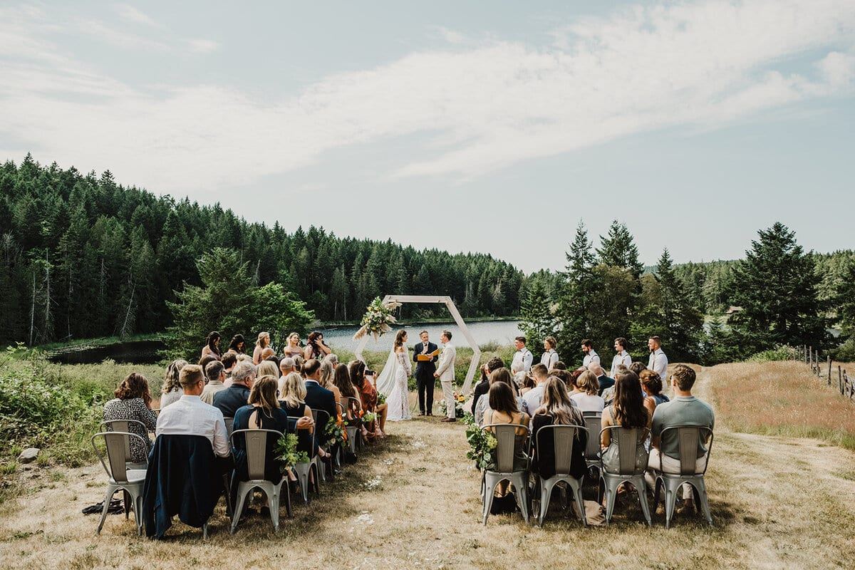 Guests watching a lakeside ceremony beneath a flower-topped arch