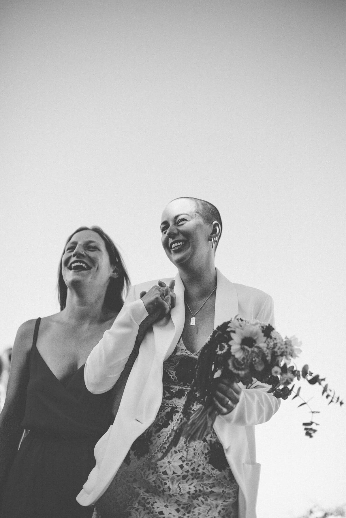 Two brides laughing together, bouquet in hand, in black and white