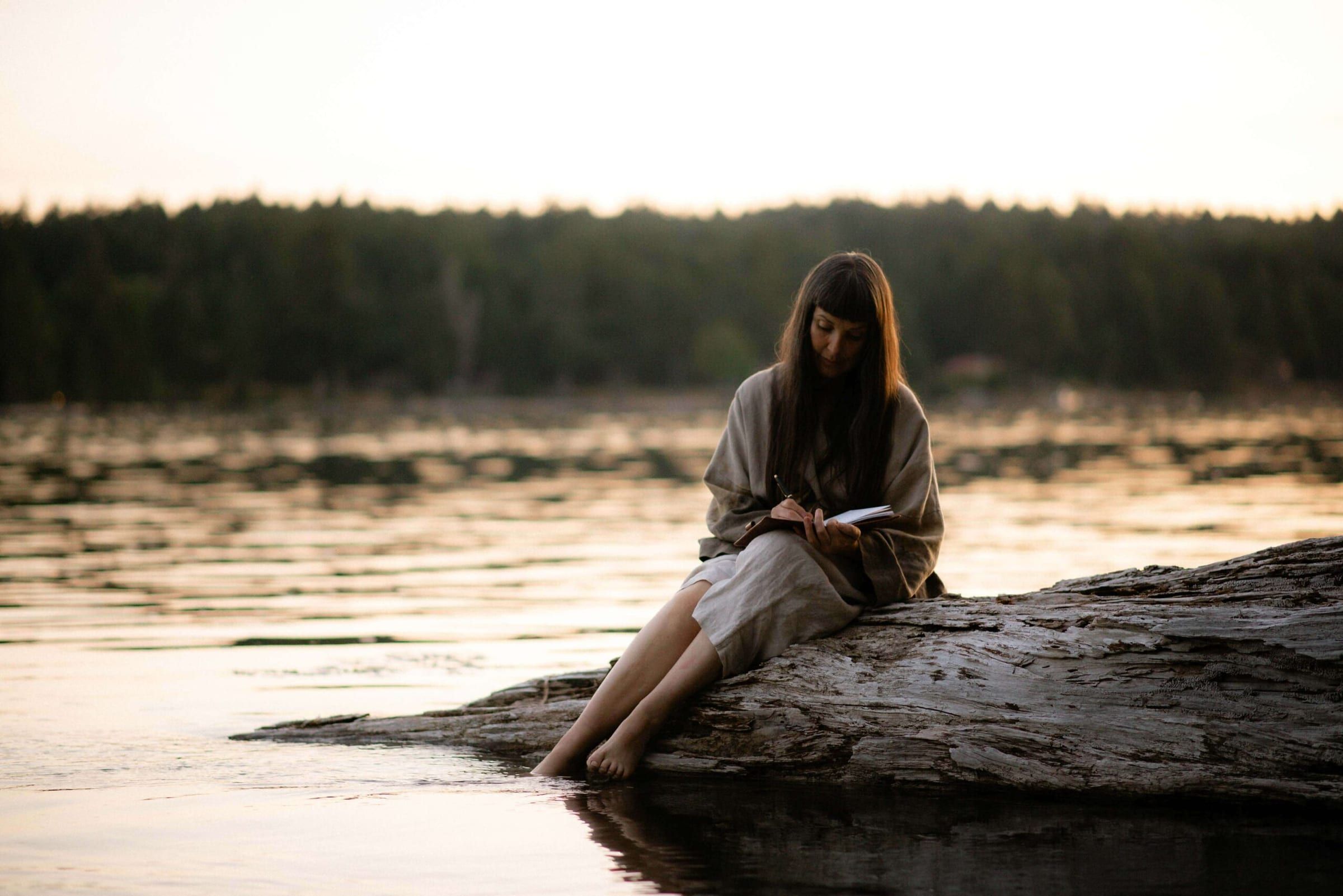 Woman sitting on a log by the water, writing in a notebook, with a serene landscape in the background.