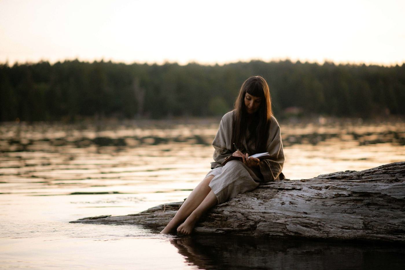 Woman sitting on a log by the water, writing in a notebook, with a serene landscape in the background.