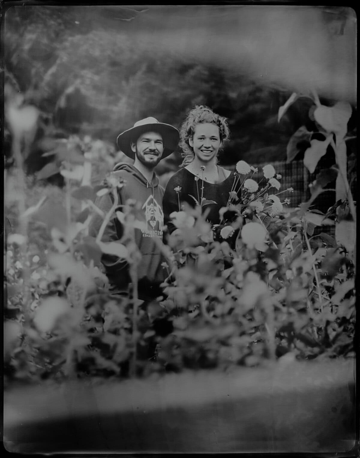 Smiling pair peeking through tall garden blooms, wet-plate style