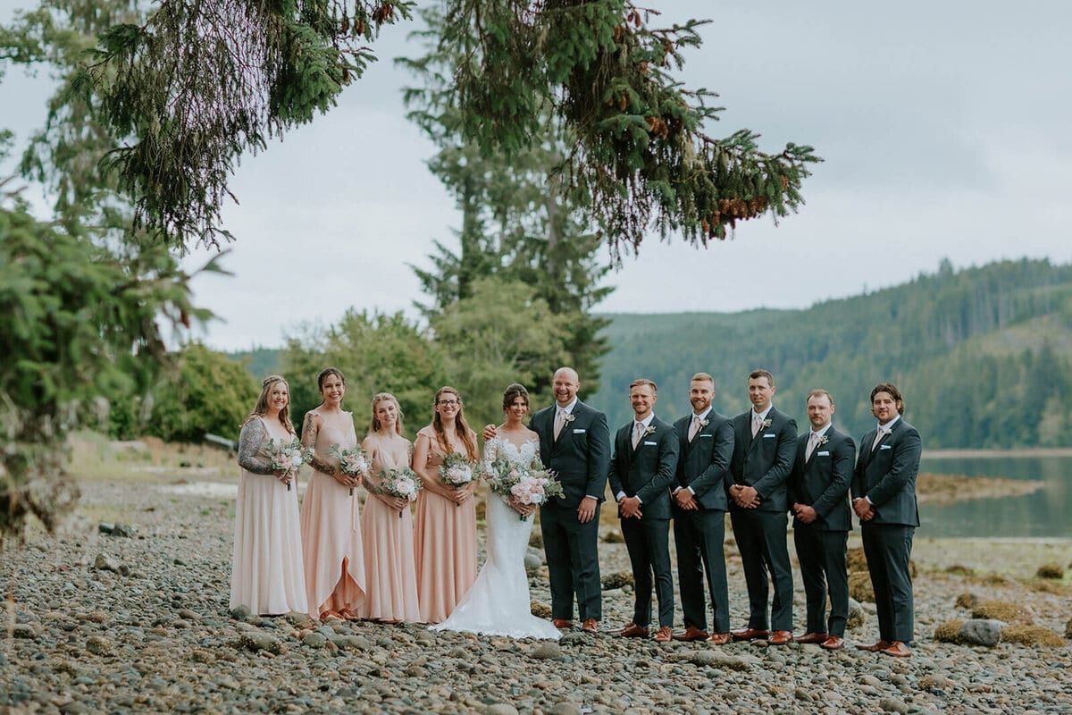 Wedding party lined up on a pebbled lakeshore beneath cedar boughs
