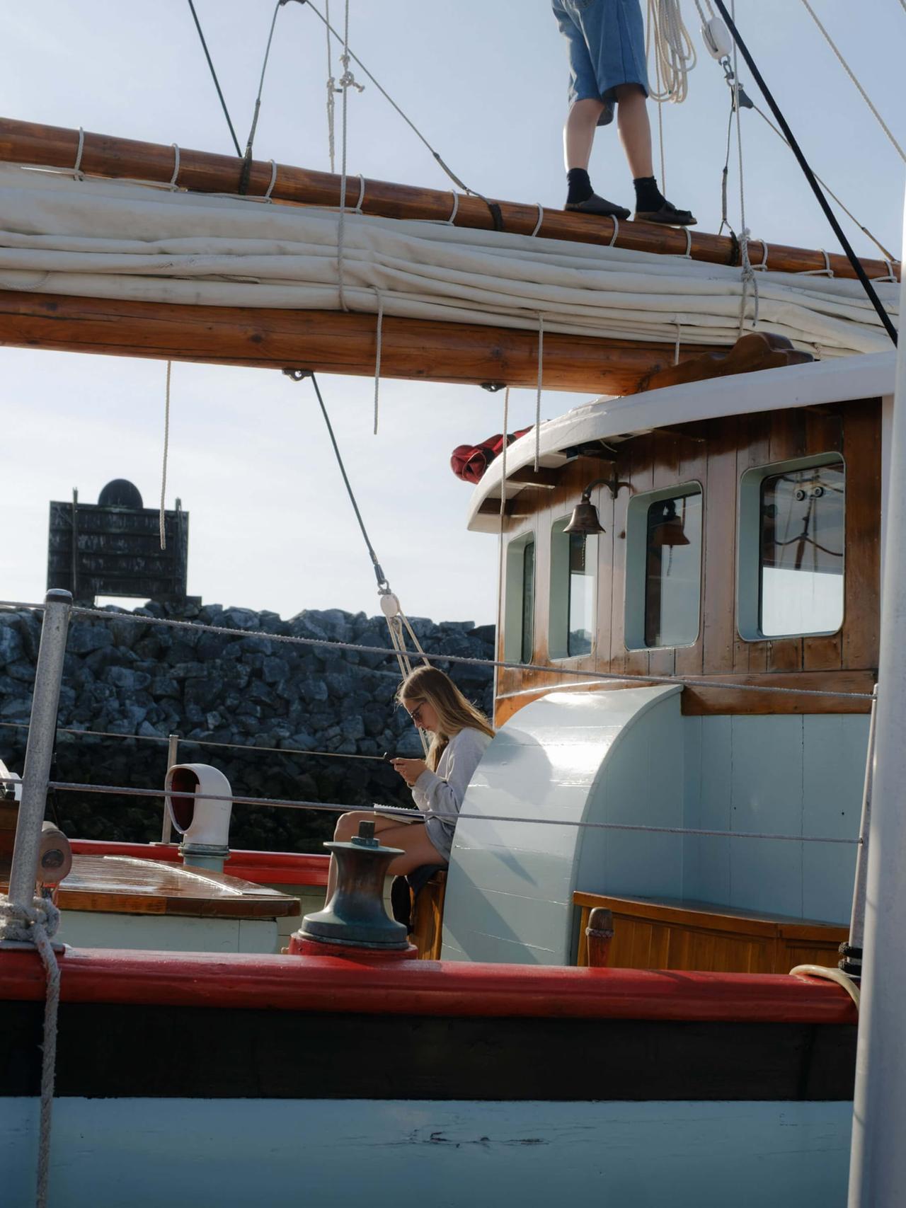 Woman sits on the deck of a wooden sailboat with rigging and boom overhead