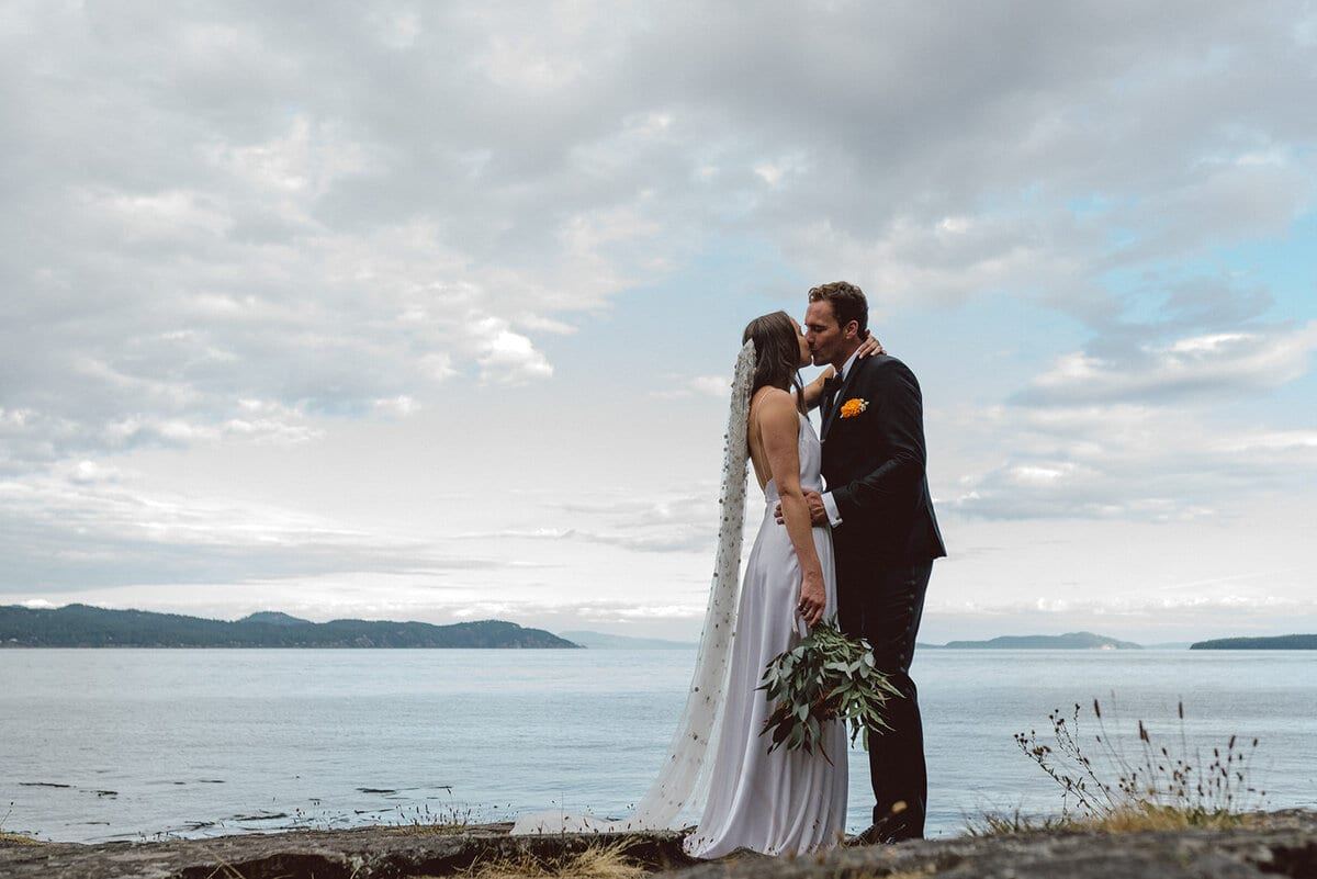 Newlyweds kissing at the ocean's edge under a wide cloudy sky