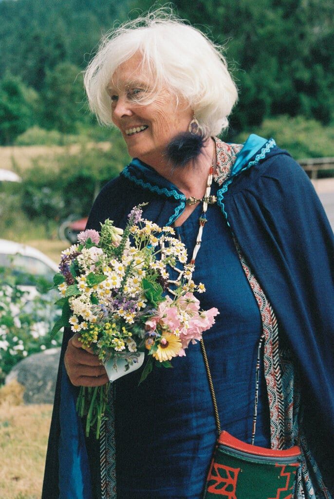 White-haired guest in an indigo cape smiling with a wildflower posy