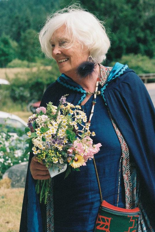 White-haired guest in an indigo cape smiling with a wildflower posy