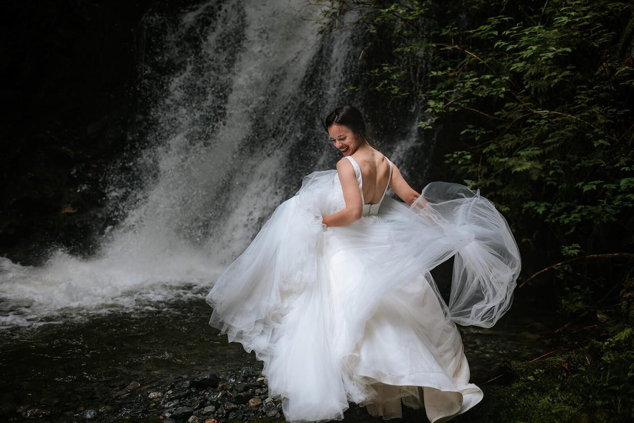 Bride in flowing white gown twirls toward weathered stone wall draped with moss at Nimmo Bay, smiling over her shoulder