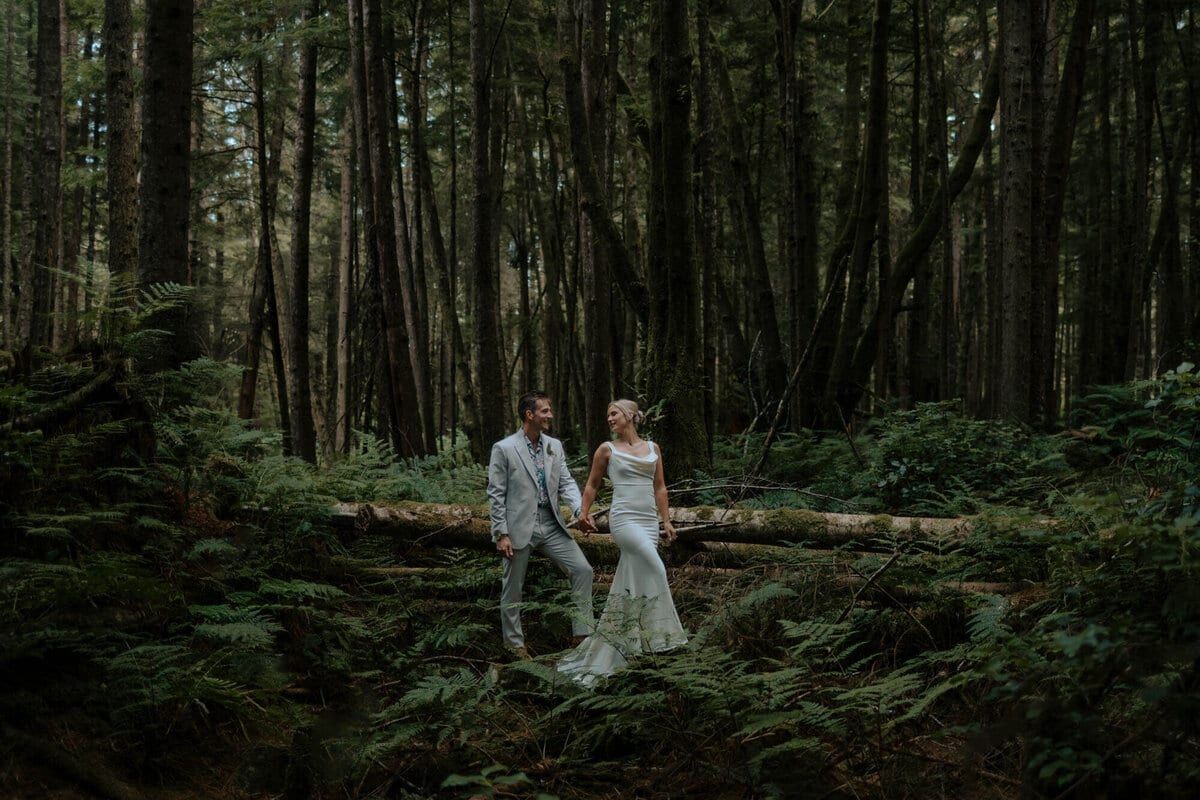 Newlyweds holding hands over fallen logs in deep mossy forest
