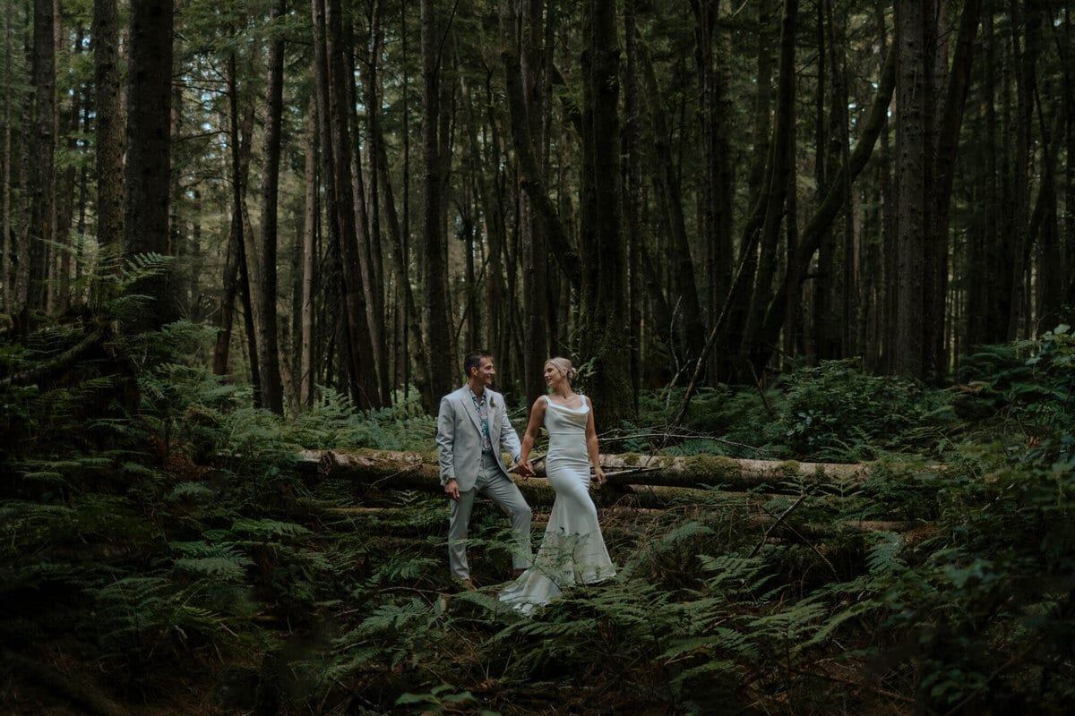 Newlyweds holding hands over fallen logs in deep mossy forest