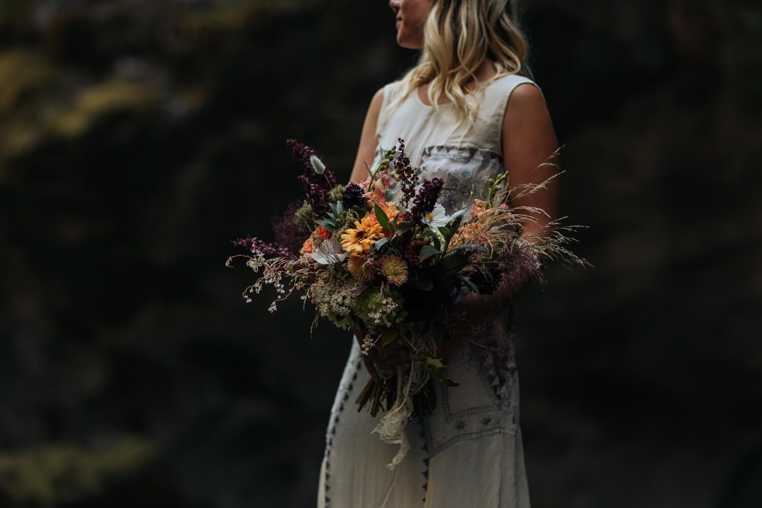 Woman holding a large bouquet of mixed flowers including orange and purple blooms, with greenery and dried elements