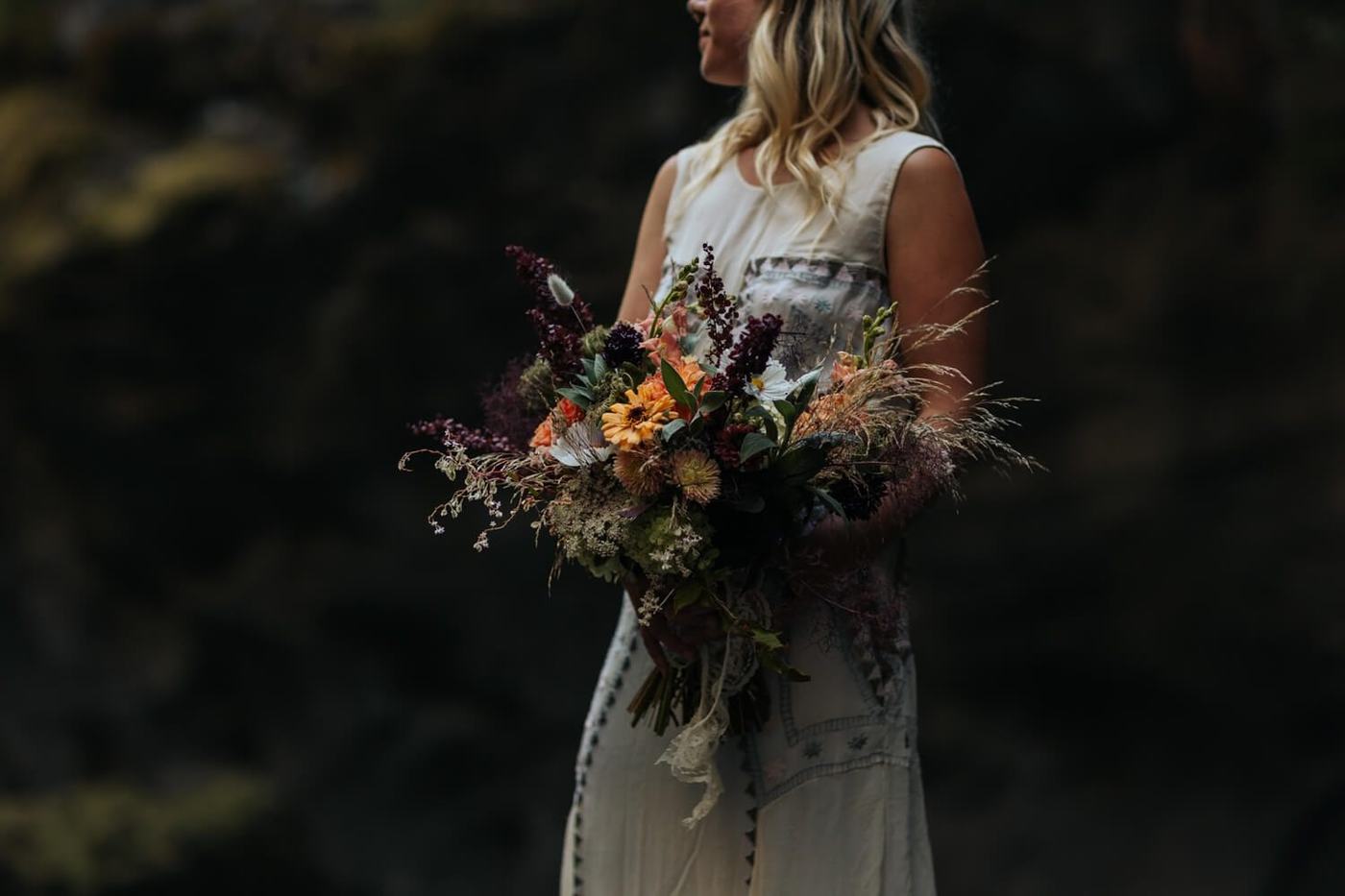 Woman holding a large bouquet of mixed flowers including orange and purple blooms, with greenery and dried elements