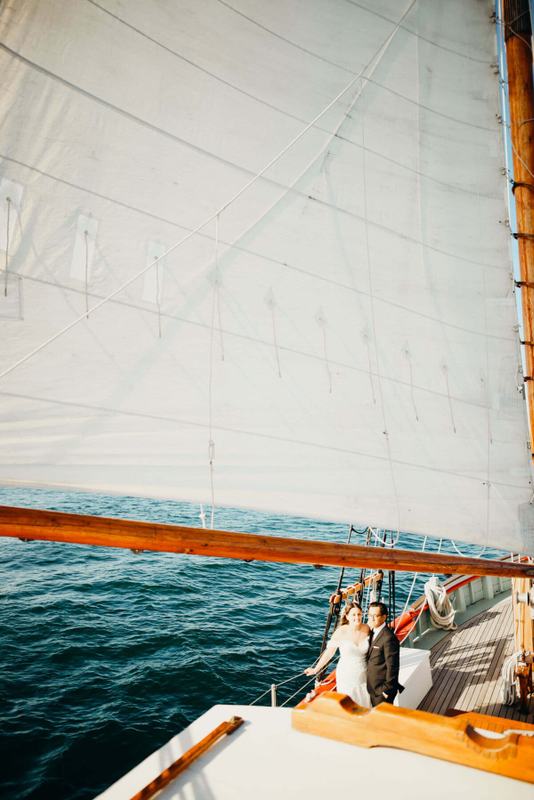 Newlyweds on deck seen past a sunlit sail over dark water
