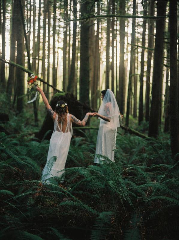 Two brides holding hands wading through ferns beneath towering trees
