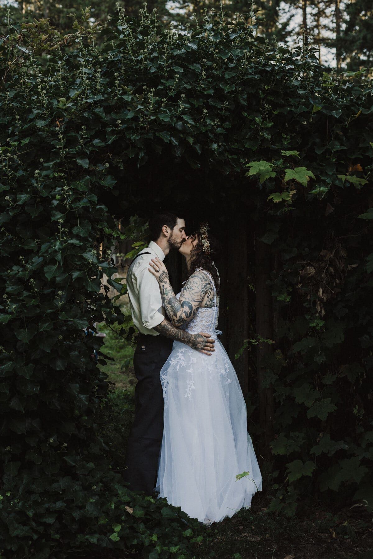 Tattooed bride and groom kissing beneath a leafy garden archway