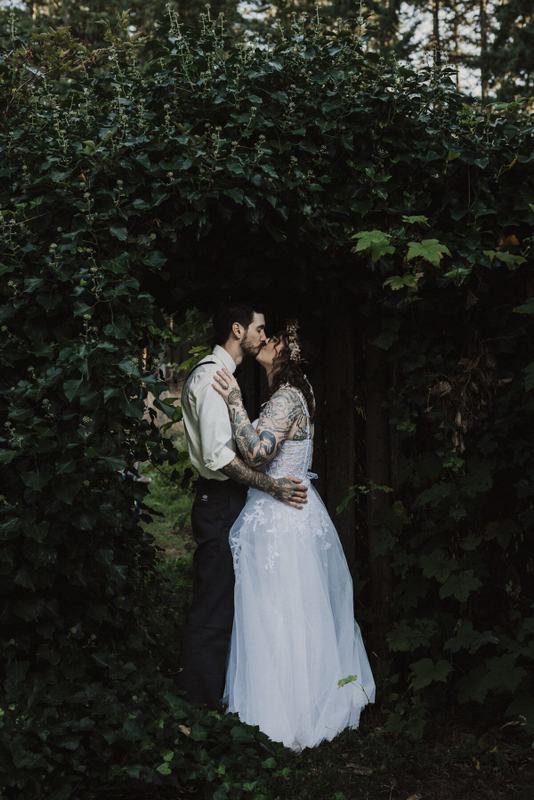 Tattooed bride and groom kissing beneath a leafy garden archway