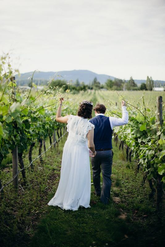 Newlyweds trailing hands along leafy vineyard rows