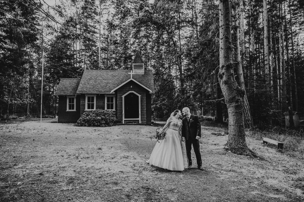 Black-and-white kiss outside a tiny chapel among tall firs