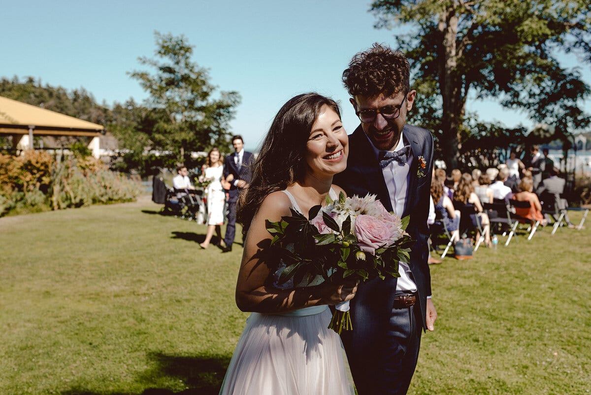 Laughing newlyweds heading up the sunny aisle with a peony bouquet