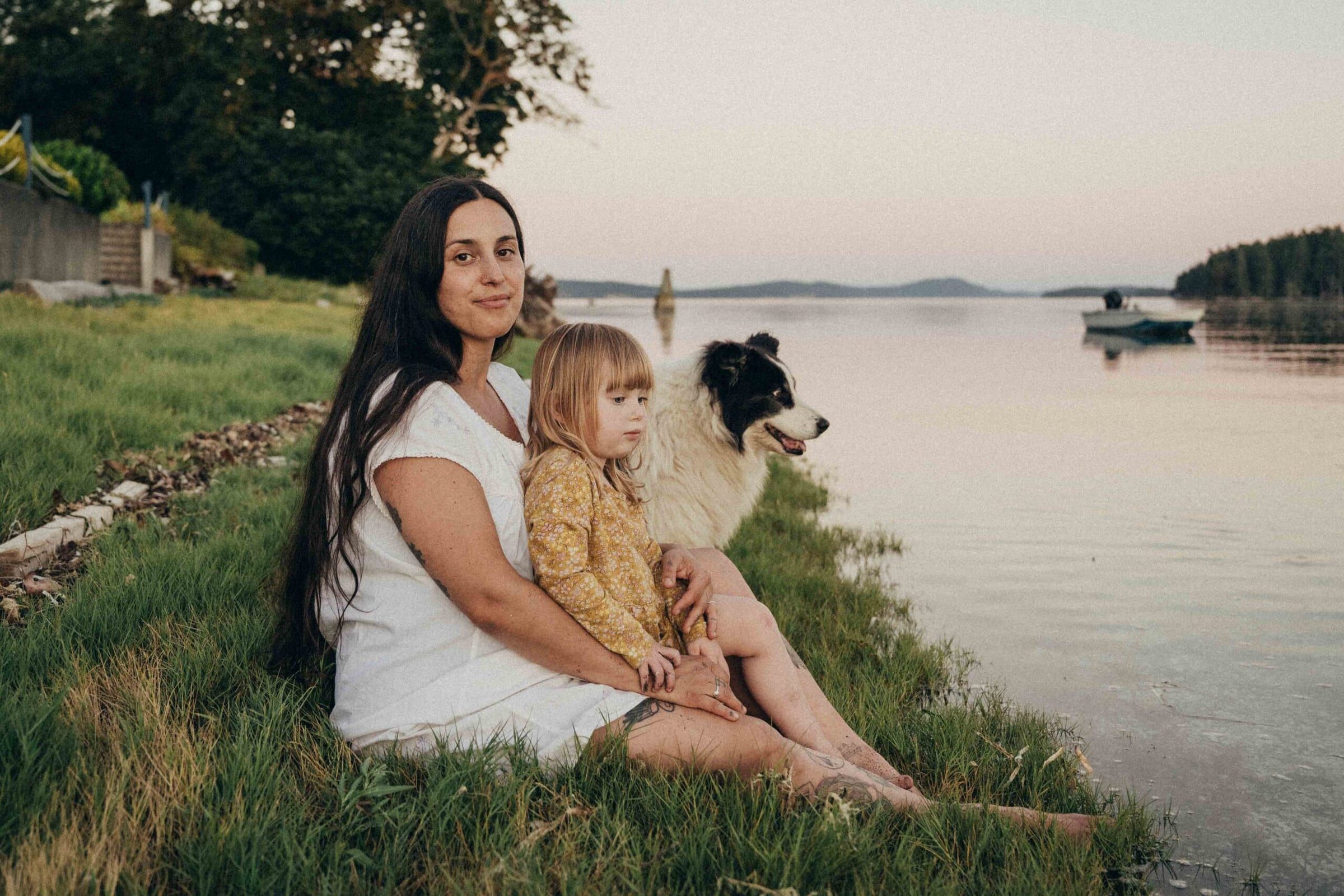 Woman with long dark hair sitting on grass by the water with a child and a dog, both looking towards the water at sunset.