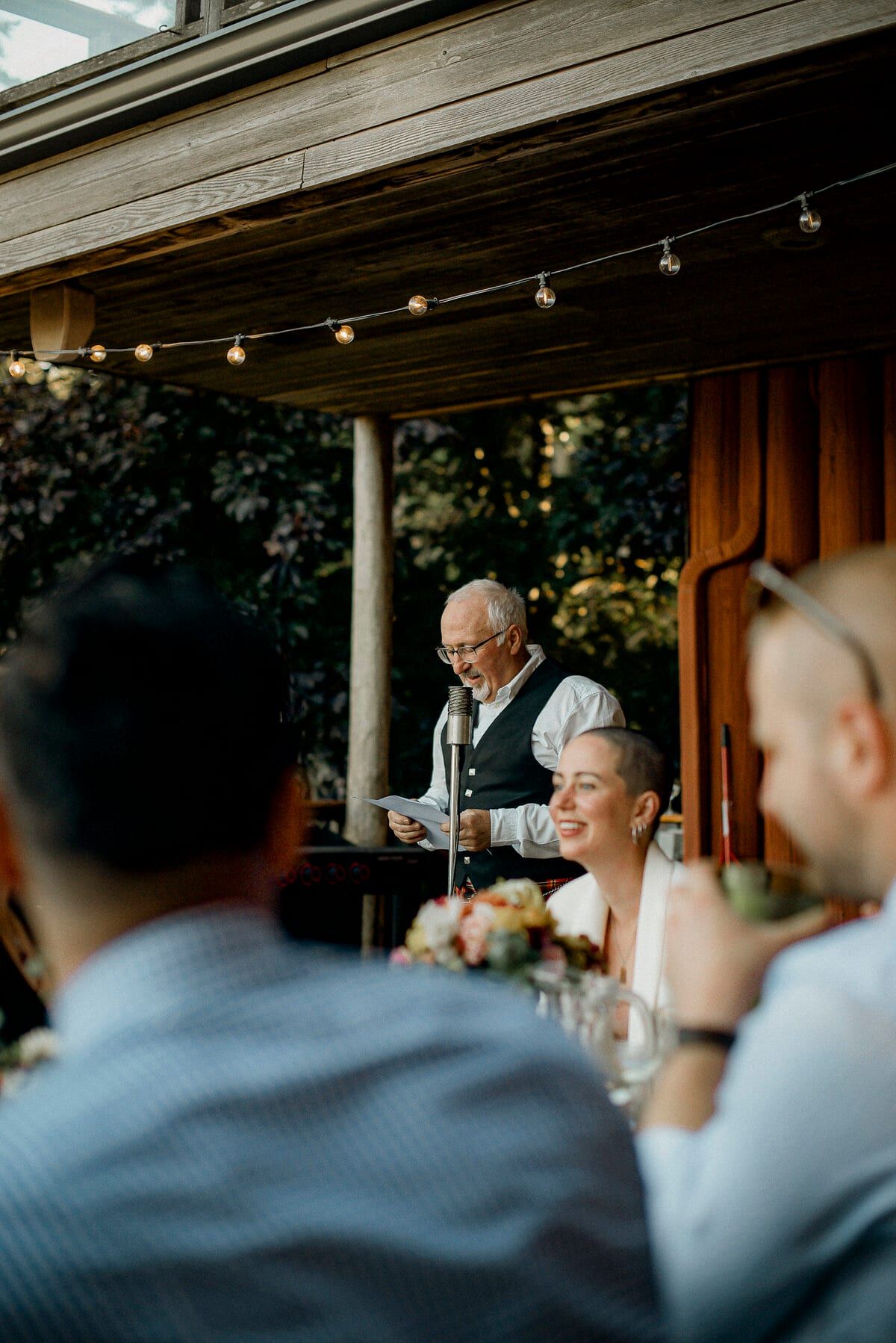 Older guest reading a speech under string lights as bride laughs