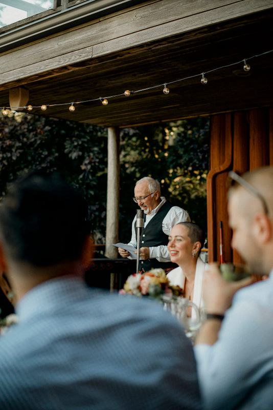 Older guest reading a speech under string lights as bride laughs