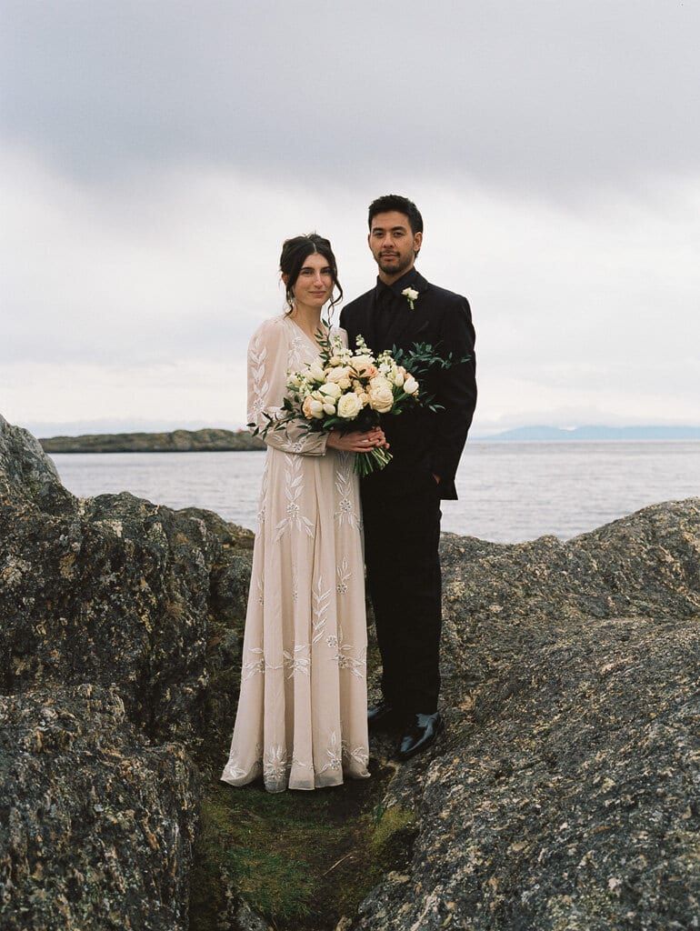 Bride and groom with white roses standing on coastal rocks under grey skies